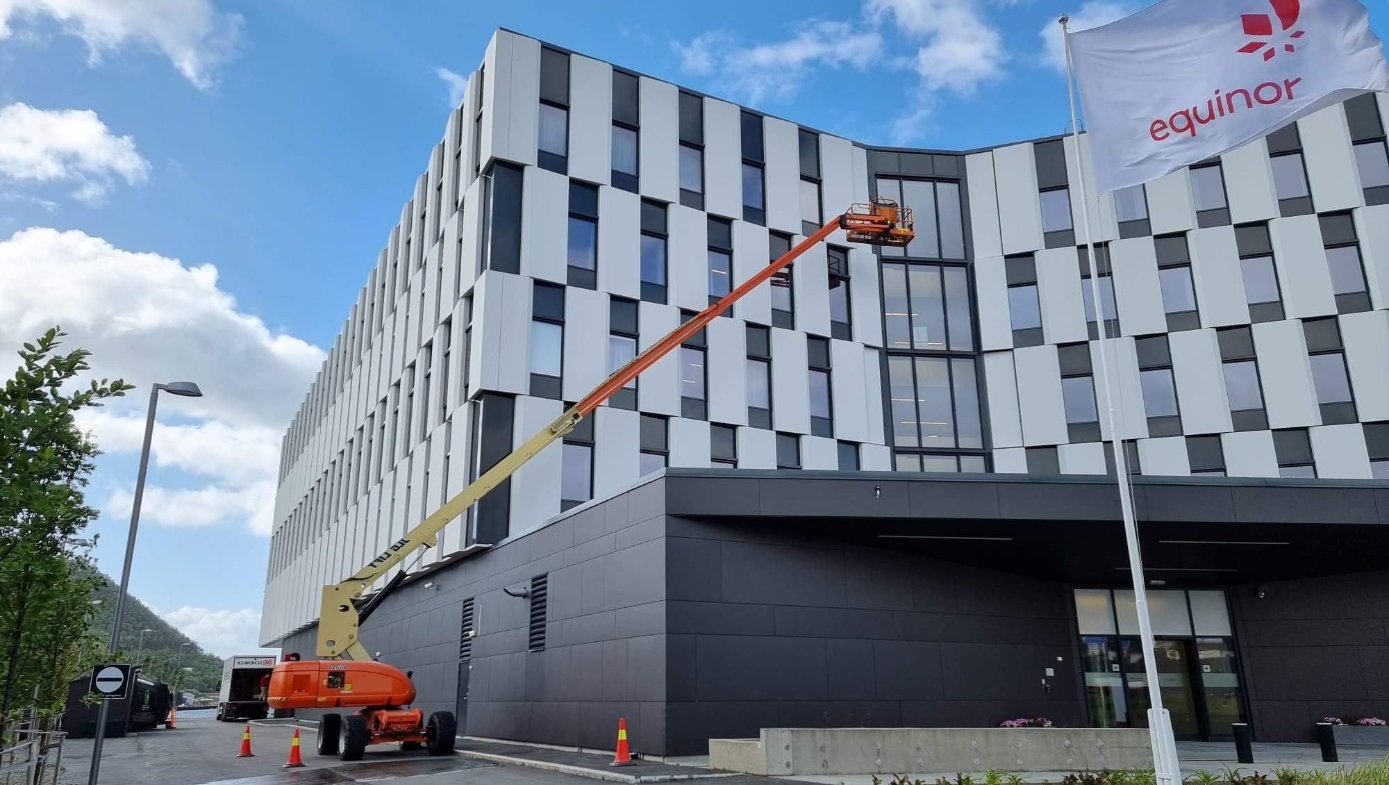Bomlift i bruk ved stort næringsbygg med Equinor-flagg. Arbeider utfører arbeid på øverste vindu.