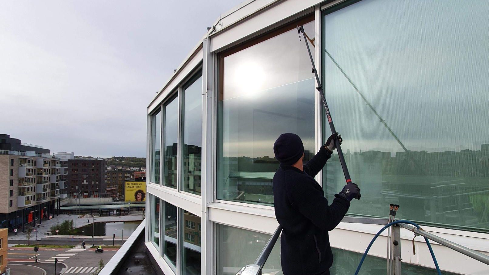 Vindusvasker står i liftkurv og rengjør store fasadevinduer i høyden med teleskopstang.