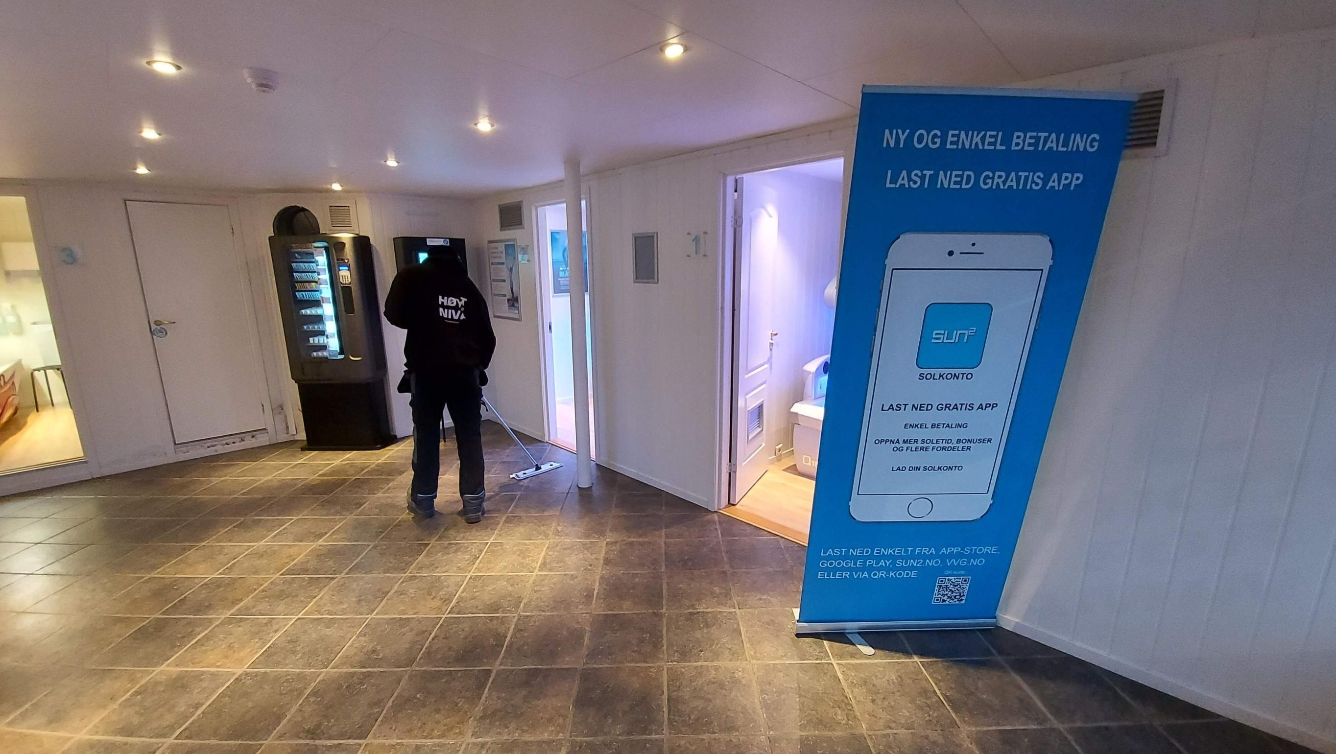 Ansatt fra Høyt Nivå utfører gulvvask i fellesområde med automater og informasjonsskilt, som del av rengjøringstjenester innendørs.